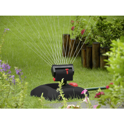Rasensprenger bewässert grünen Garten; Wasserstrahlen formen gleichmäßigen Halbkreis. Blumen und Pflanzen im Hintergrund.